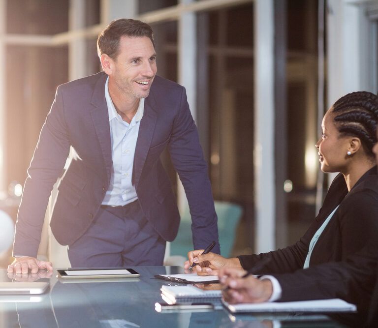A man in a blue suit stands and smiles while speaking to two colleagues seated at a conference table with notebooks and a tablet in a modern office setting.