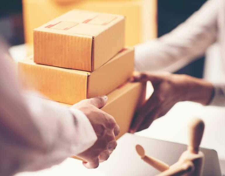 Two people exchange a stack of three small cardboard boxes, suggesting a delivery or package handoff. A wooden mannequin figure is visible on the table in the foreground.