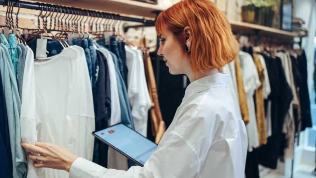 A person with short red hair wearing a white shirt uses a tablet while inspecting clothes hanging on racks in a clothing store.