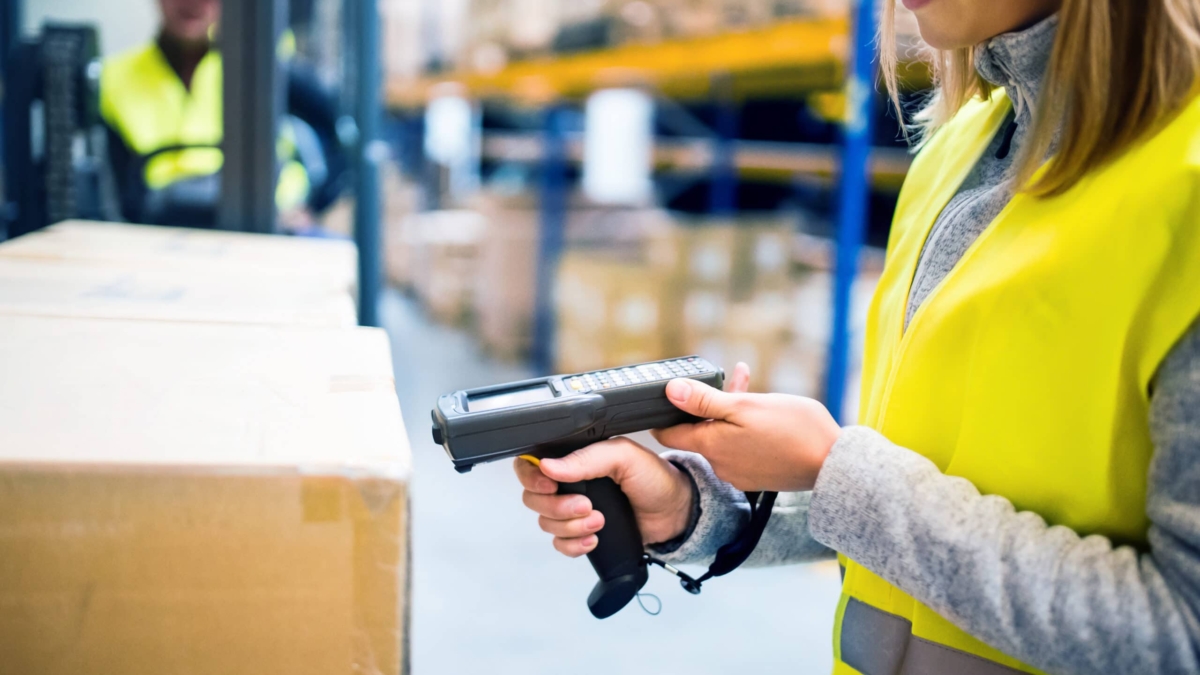 A worker in a yellow safety vest scans a cardboard box with a handheld barcode scanner in a warehouse. Shelves with more boxes and another worker in the background are visible.
