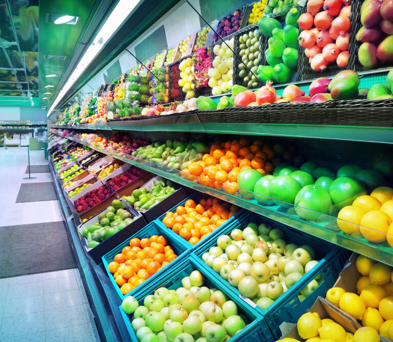 Shelves in a grocery store filled with a colorful variety of fresh fruits and vegetables, including apples, oranges, lemons, and peppers, neatly arranged in rows and baskets.