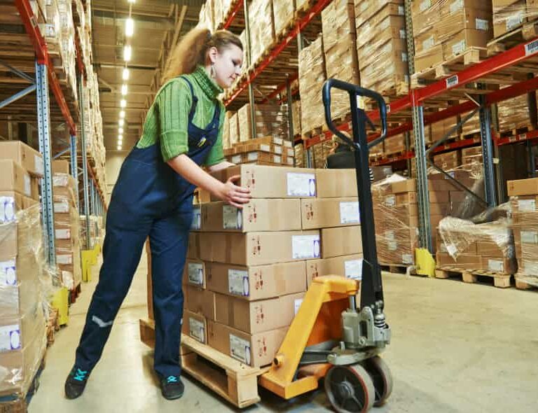 A woman in work overalls moves a pallet stacked with cardboard boxes using a pallet jack in a warehouse filled with shelves of boxed goods.