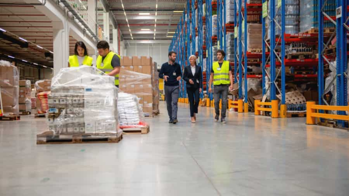 Four people in safety vests walk through a spacious warehouse with shelves stacked high with boxes and goods, while two workers handle wrapped pallets in the foreground.