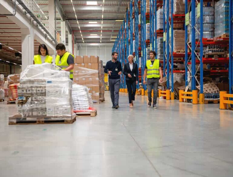 Four people in safety vests walk through a spacious warehouse with shelves stacked high with boxes and goods, while two workers handle wrapped pallets in the foreground.