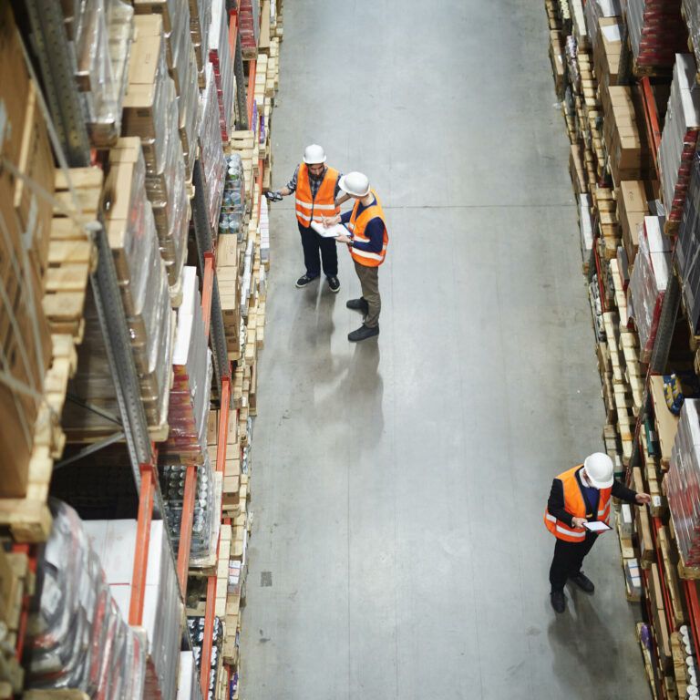 Three workers in orange safety vests and white helmets check inventory on high shelves in a large warehouse, viewed from above. The aisles are lined with stacked boxes and pallets.