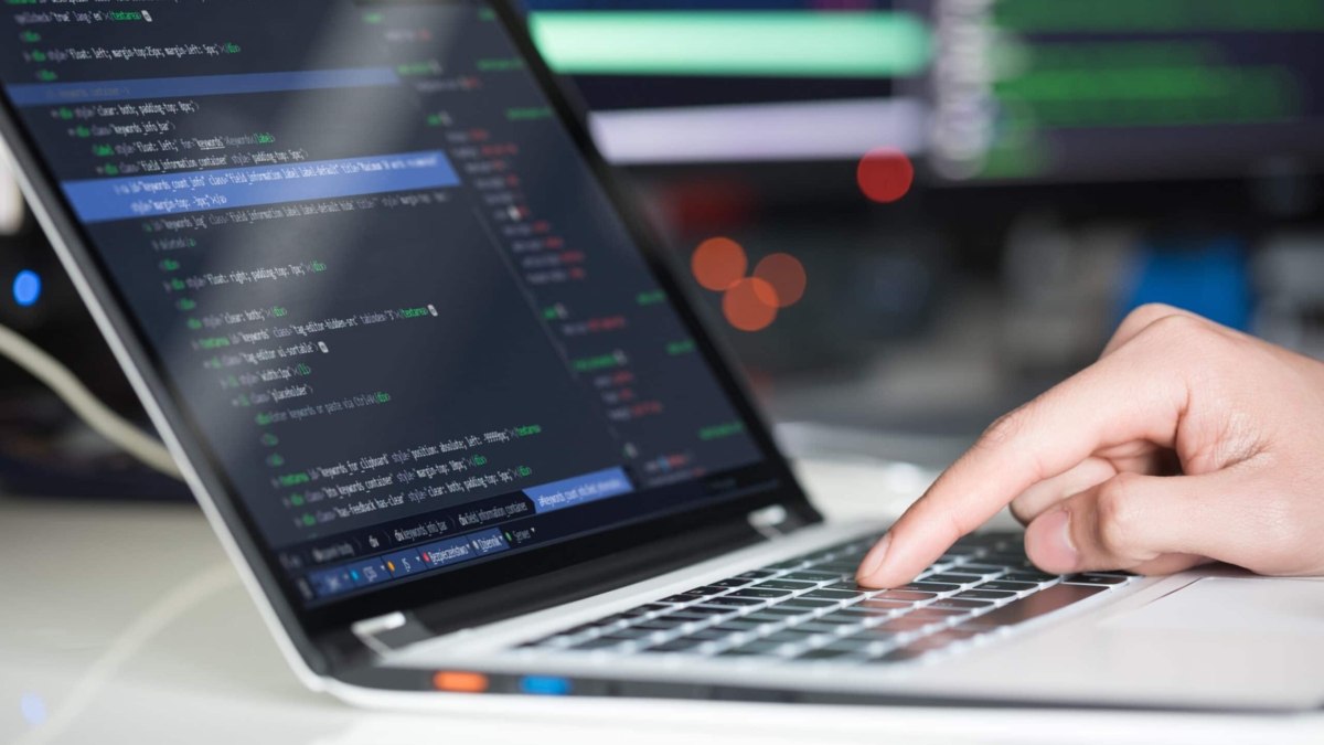 A hand types on a laptop keyboard, with the screen displaying lines of code in a programming environment. The background is blurred, showing another monitor with code.