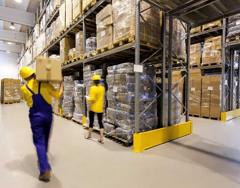 Workers in yellow safety helmets and uniforms move boxes in a large warehouse filled with stacked pallets, shelves, and packaged goods, with high ceilings and bright lighting.