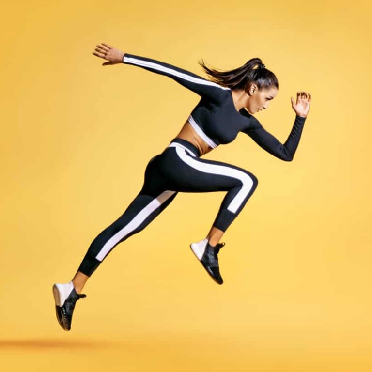 Woman in black and white athletic wear jumps in mid-air against a solid yellow background, appearing energetic and focused, with her arms and legs extended in a running pose.