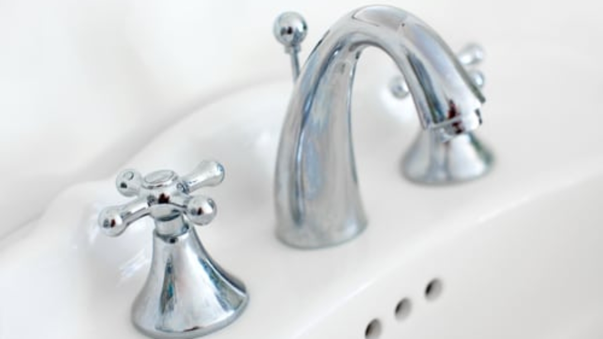 Close-up of a shiny chrome bathroom faucet with traditional cross-shaped hot and cold handles on a white ceramic sink. Three small overflow drain holes are visible below the faucet.