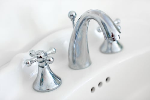 Close-up of a shiny chrome bathroom faucet with traditional cross-shaped hot and cold handles on a white ceramic sink. Three small overflow drain holes are visible below the faucet.