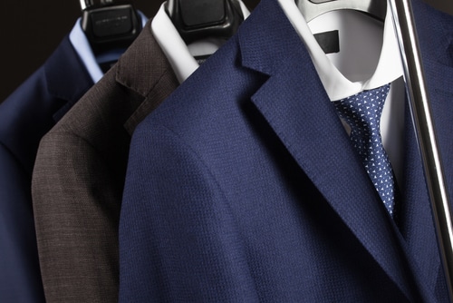 Three suits on hangers, including a blue suit with a white shirt and blue polka dot tie in front, and two darker suits behind it, all hanging on a clothing rack.