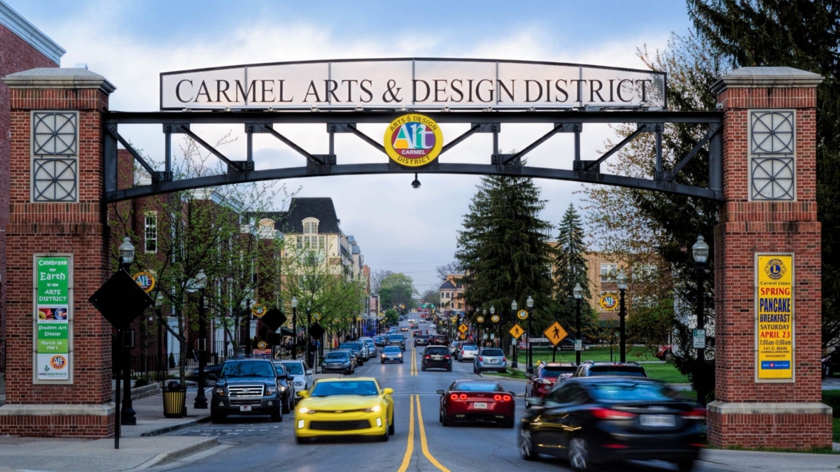 A street scene in Carmel Arts & Design District, Indiana, with a large archway sign overhead, cars driving on the road, trees, buildings, and people walking on the sidewalks. A yellow car is prominent in the foreground.