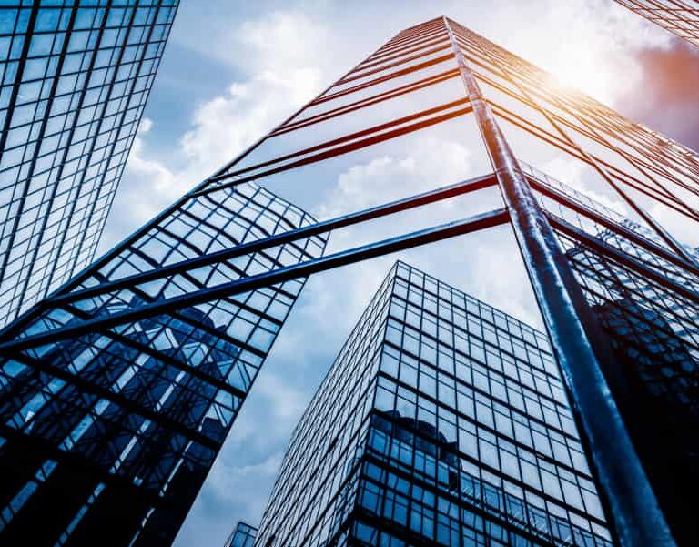 Tall glass skyscrapers reflect the blue sky and clouds as sunlight streams between the buildings, creating an urban cityscape with a modern architectural feel.