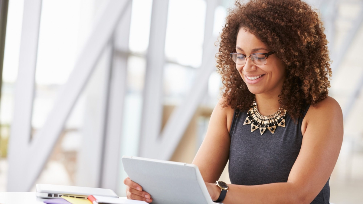 A woman with curly hair and glasses sits at a desk, smiling while looking at a tablet. Papers, a notebook, and a laptop with power platform dashboards open are on the desk in front of her. The background shows a bright, modern interior.