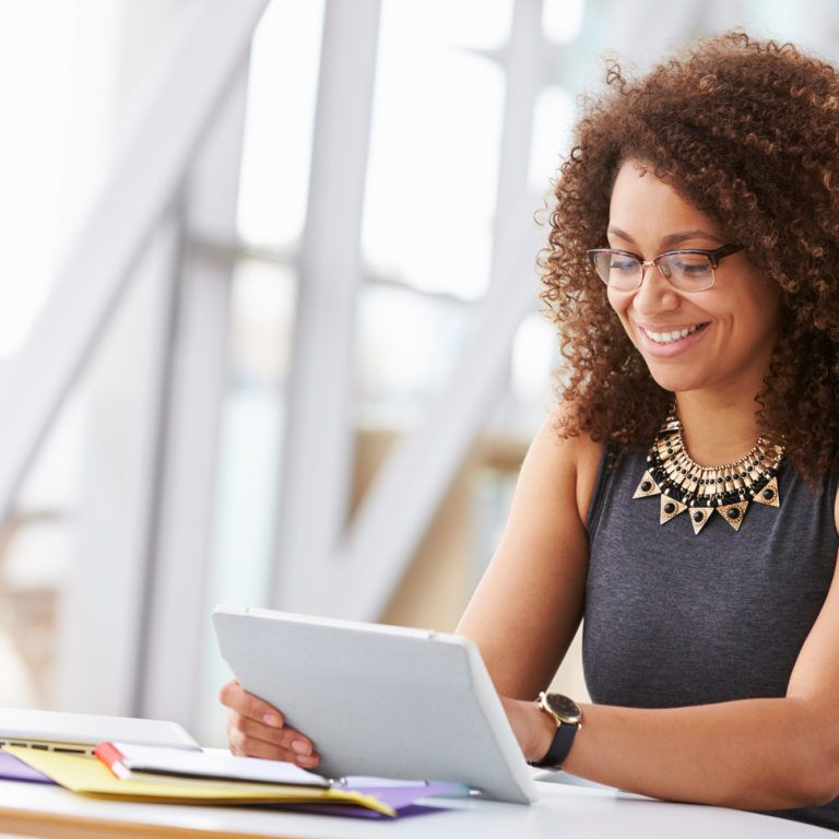 A woman with curly hair and glasses sits at a desk, smiling while looking at a tablet. Papers, a notebook, and a laptop with power platform dashboards open are on the desk in front of her. The background shows a bright, modern interior.