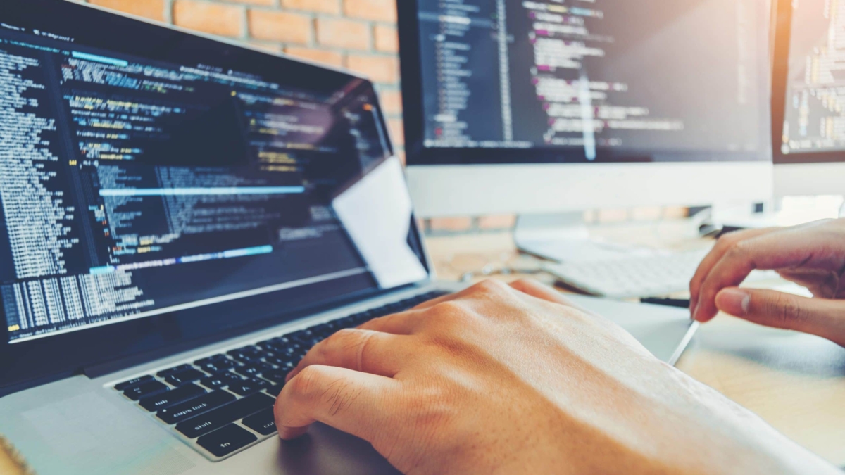 A person types on a laptop with programming code visible on the screen, possibly working on enVista Careers or a parcel audit service. Another monitor in the bright workspace, set against a brick wall, also displays code.
