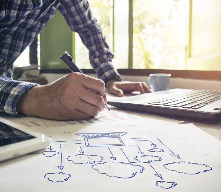 A person in a plaid shirt writes on paper next to a laptop, tablet, and a cloud computing diagram featuring D365 Business Central, with sunlight streaming through a window in the background.