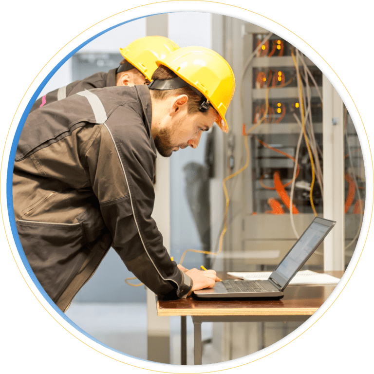 Two workers wearing yellow hard hats and dark jackets lean over a laptop on a desk, utilizing Dynamics 365 Field Service, with network equipment and cables visible in the background.