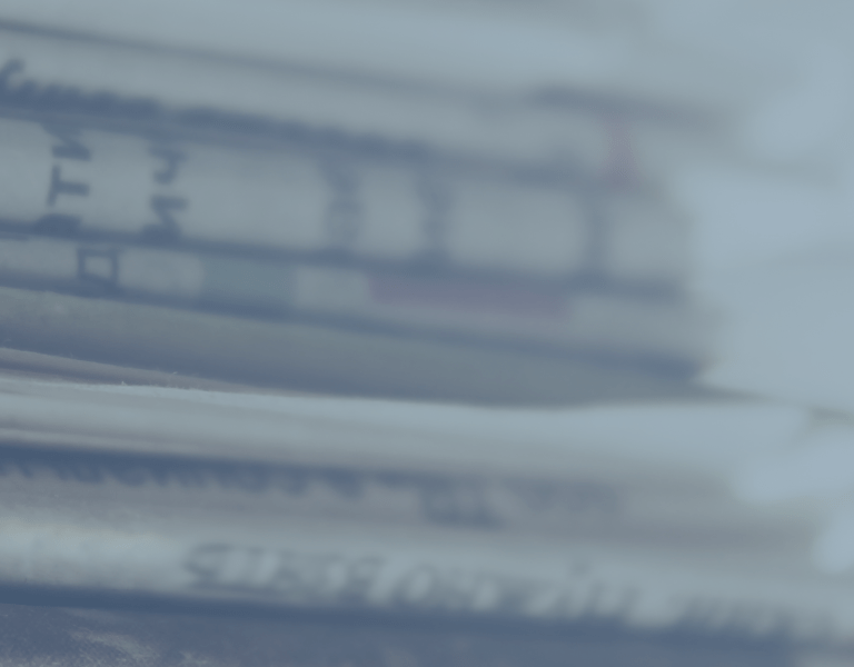 A close-up view of a stack of newspapers, with printed text visible on the pages. The image has a blue tint and a blurred background.