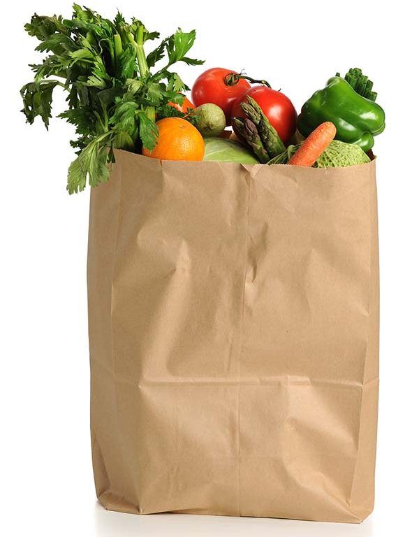 A brown paper grocery bag filled with fresh vegetables and fruits, including celery, tomatoes, green bell pepper, carrots, an orange, and asparagus—ready for transportation network optimization—against a white background.