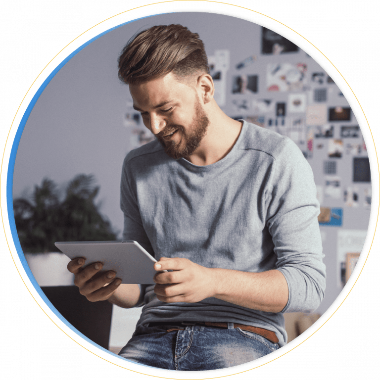 A smiling man with a beard, in a light gray long-sleeve shirt and jeans, sits indoors looking at a tablet, possibly exploring Microsoft Dynamics 365 Project Operations. A blurred collage of photos and a plant appear in the background.
