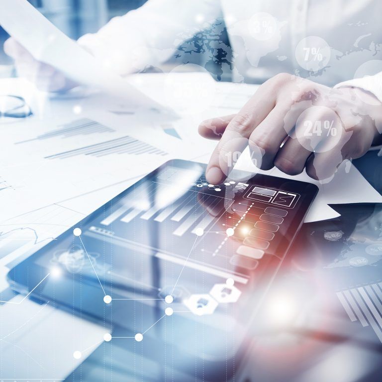 A person reviews financial reports and graphs on paper and a tablet, with data and analytics icons overlaid, suggesting business analysis enhanced by innovative technology solutions.