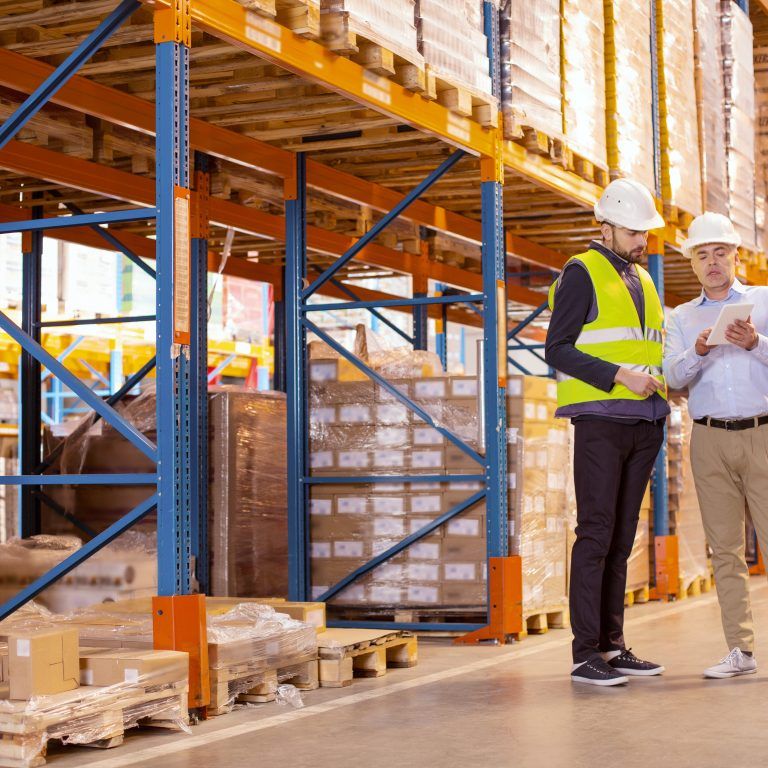 Two men in safety gear stand in a warehouse aisle lined with shelves and boxes. One wears a reflective vest and hard hat; the other holds a tablet, likely reviewing inventory using a labor management system as they discuss logistics.