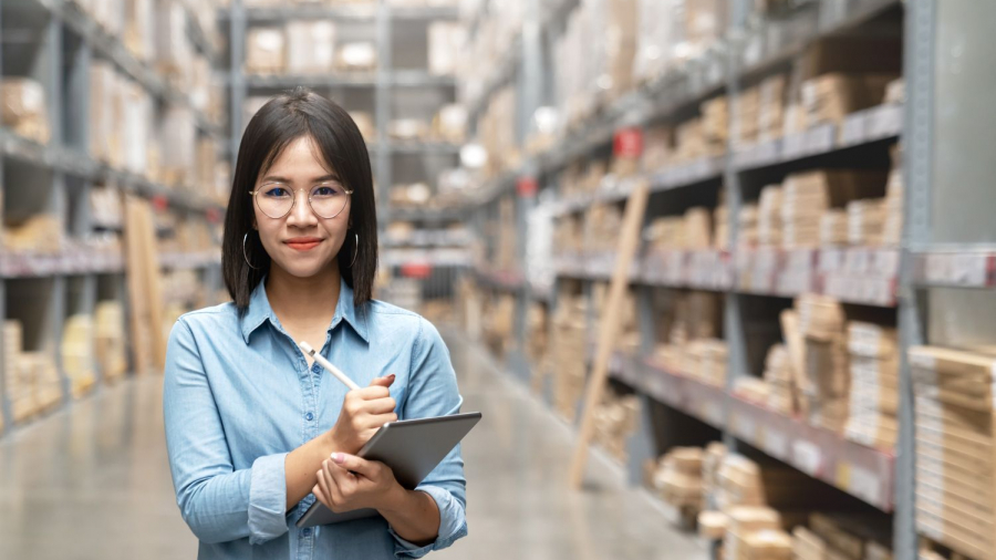 A woman with glasses and a blue shirt stands in a warehouse, holding a clipboard and pen, smiling at the camera. Shelves filled with boxes and supplies suggest efficient inventory management with Dynamics 365 Business Central.