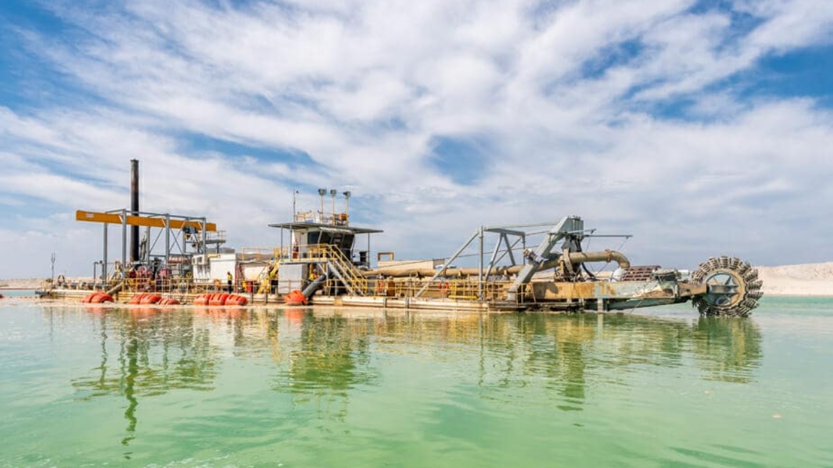 A large industrial dredging machine floats on green water under a partly cloudy blue sky, with pipes and equipment visible on its deck.