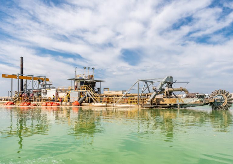 A large industrial dredging machine floats on green water under a partly cloudy blue sky, with pipes and equipment visible on its deck.
