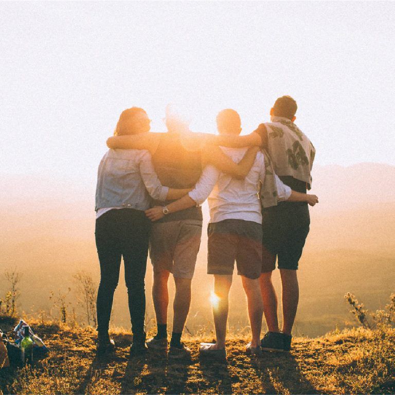 Four people stand side by side with arms around each other, looking at a sunset over a scenic, hilly landscape. The warm sunlight creates an envista of golden glow around them.