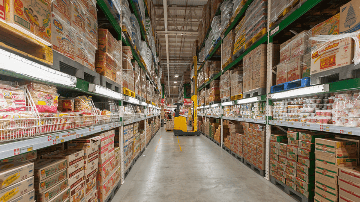 Warehouse aisle filled with stacked shelves of packaged goods, primarily instant noodles and food products; a worker operates a yellow pallet jack in the distance under bright overhead lighting.