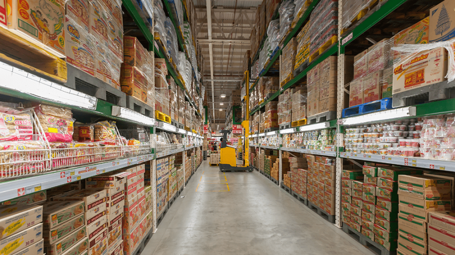 Warehouse aisle filled with stacked shelves of packaged goods, primarily instant noodles and food products; a worker operates a yellow pallet jack in the distance under bright overhead lighting.
