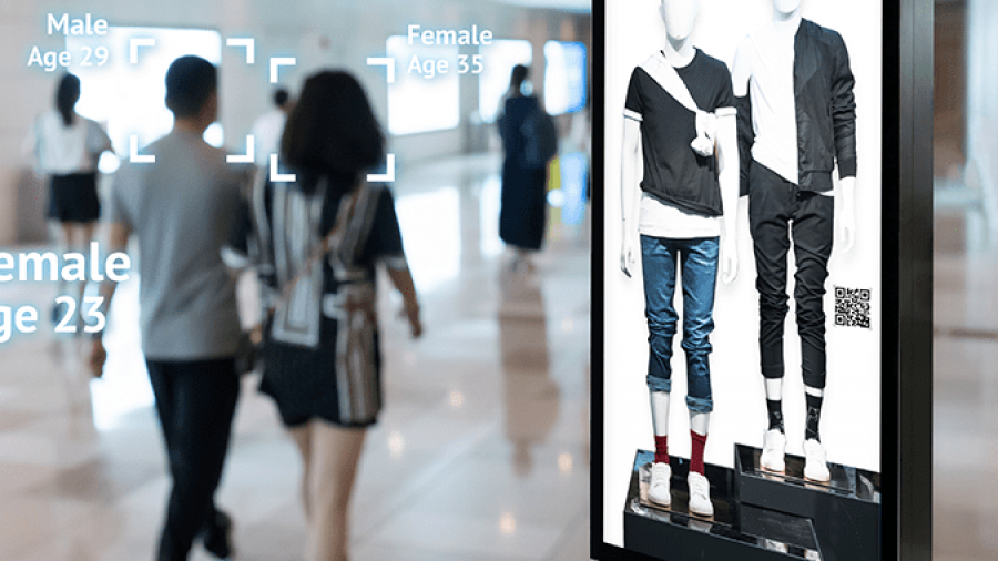A digital billboard in a mall displays mannequins in casual clothing. Nearby, a group of people walks past, while facial recognition technology identifies their gender and estimated age on screen overlays.