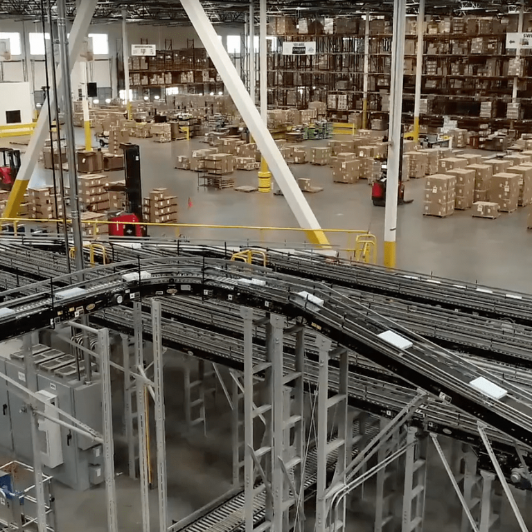 Large warehouse interior with rows of shelves stacked with boxes, several forklifts moving pallets, and a network of automated conveyor belts in the foreground. The space is well-lit and organized for distribution.