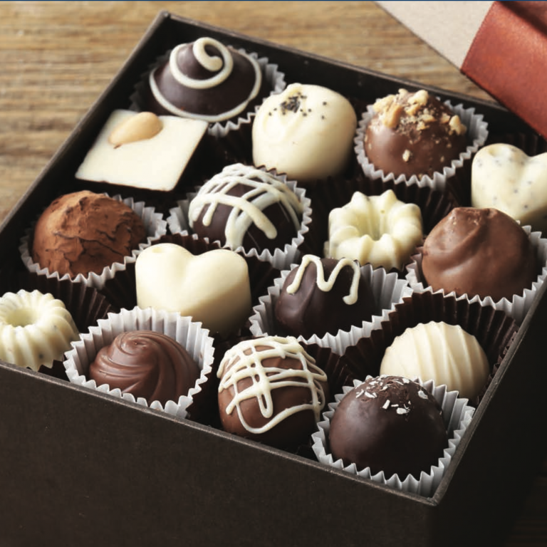 An open box of assorted chocolates, including heart-shaped, round, and square pieces in dark, milk, and white chocolate, displayed neatly in paper cups on a wooden surface.