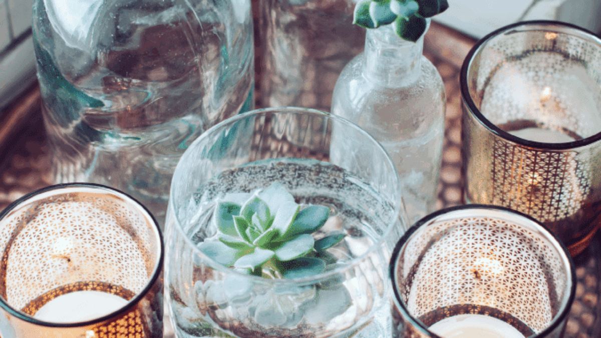 A copper tray displays three glass bottles with succulent plants, a glass bowl with a floating succulent, and two lit patterned candle holders, creating a cozy and stylish arrangement.