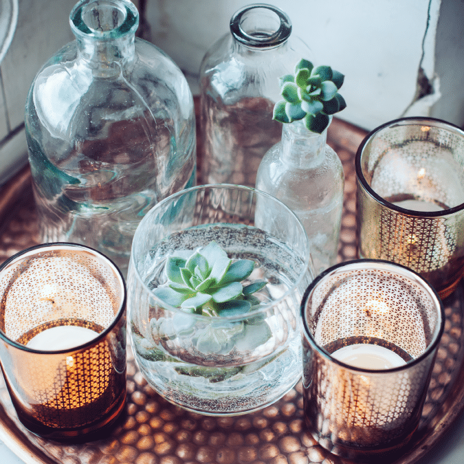 A copper tray displays three glass bottles with succulent plants, a glass bowl with a floating succulent, and two lit patterned candle holders, creating a cozy and stylish arrangement.