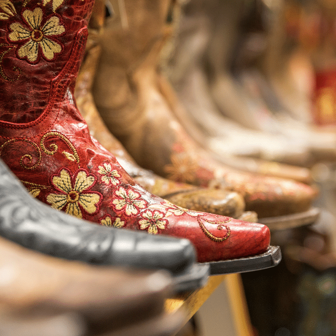 Close-up of an ornate red cowboy boot with yellow floral embroidery, displayed among several other brown and tan cowboy boots in the background on a shelf.