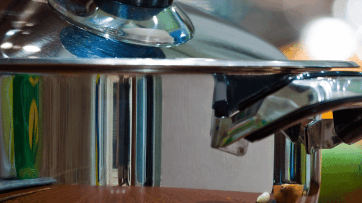 A close-up of a shiny stainless steel pot with a glass lid, sitting on a wooden surface. The pots handle and lid knob are visible, reflecting colorful shapes from the surrounding kitchen.