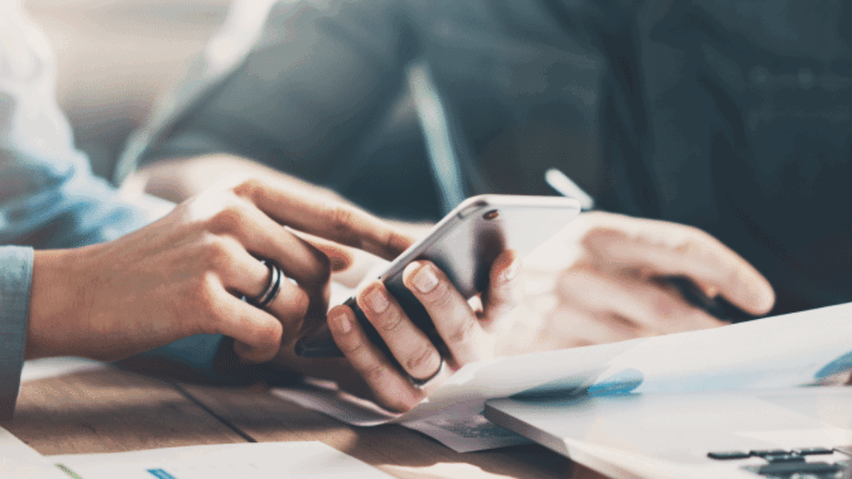 Two people sit at a desk with documents and a laptop, focusing on a smartphone as one person points at the screen, suggesting a discussion or collaboration on work or data.