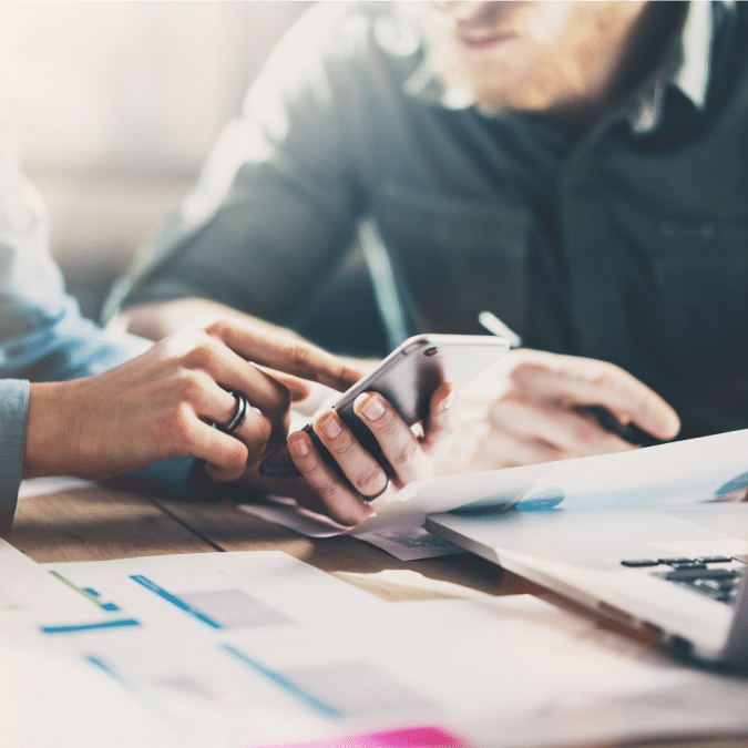 Two people sit at a desk with documents and a laptop, focusing on a smartphone as one person points at the screen, suggesting a discussion or collaboration on work or data.