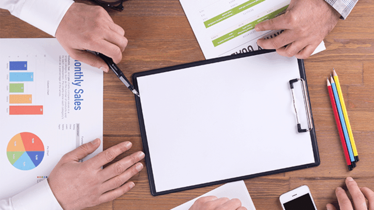 Four people sit around a wooden table with documents, charts, a blank clipboard, colored pencils, a smartphone, eyeglasses, and a coffee cup, suggesting a business meeting or discussion.