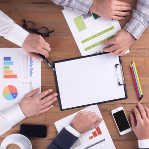 Four people sit around a wooden table with documents, charts, a blank clipboard, colored pencils, a smartphone, eyeglasses, and a coffee cup, suggesting a business meeting or discussion.