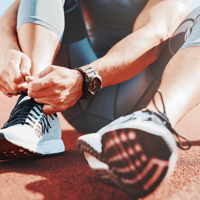 Close-up of a person in athletic clothing sitting on a red running track, tying the laces of their white and black running shoes. Sunlight highlights their arm and wristwatch.
