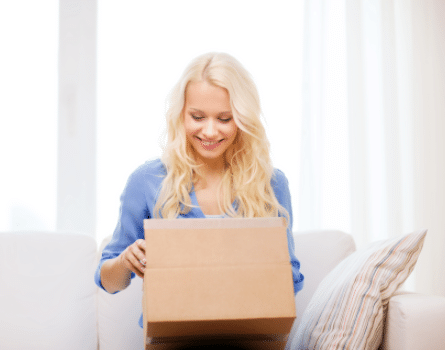 A smiling woman with long blonde hair sits on a couch and opens a cardboard box, looking inside with excitement. A striped pillow is beside her, and soft daylight fills the room.