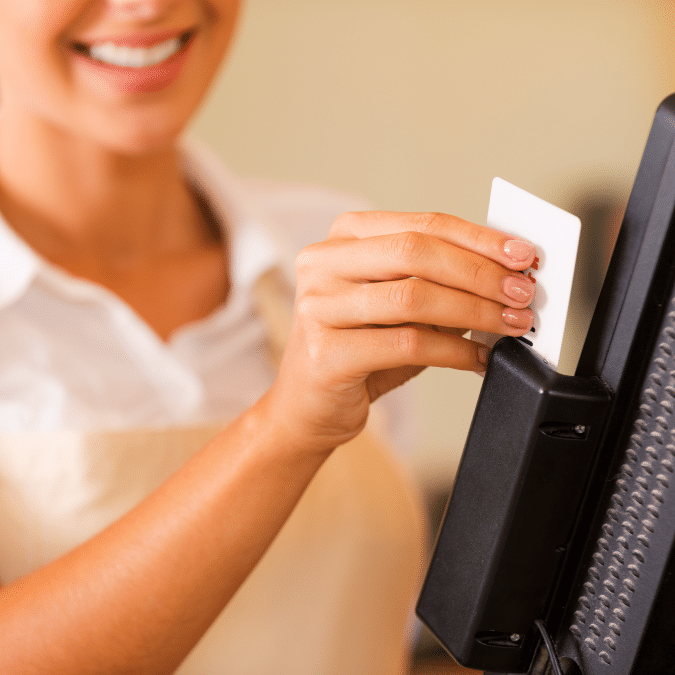 A smiling person in an apron swipes a white card through a card reader attached to a point-of-sale terminal. Only the lower half of their face and upper body are visible.