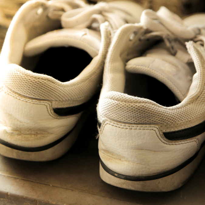 A close-up of the back of a pair of worn white sneakers with dark stripes, resting side by side on a surface. The shoes show signs of use, with creases and slight dirt marks visible.