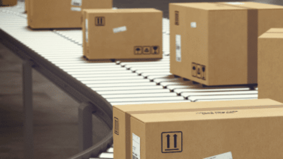 Several cardboard boxes move along a conveyor belt in a warehouse or shipping facility, with shipping labels and handling symbols visible on the packages.