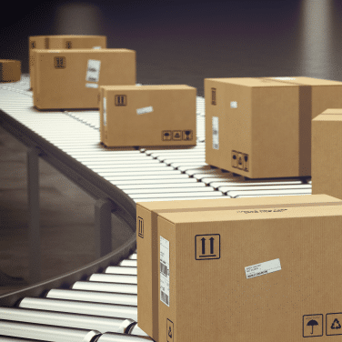 Several cardboard boxes move along a conveyor belt in a warehouse or shipping facility, with shipping labels and handling symbols visible on the packages.
